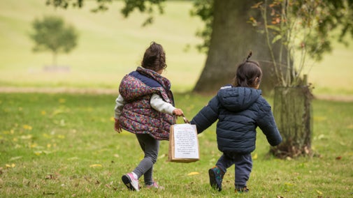 Two small Asian children in coats walking in parkland with tree in background